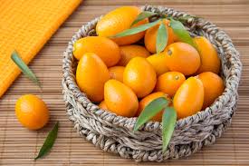 A woven basket filled with small, bright orange kumquat citrus fruits, some with green leaves, placed on a bamboo mat with an orange cloth and a few loose kumquats and leaves beside it.
