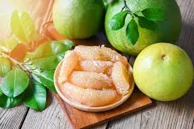 A wooden surface displaying a small plate of peeled pomelo segments, surrounded by whole pomelos with green skins and fresh green leaves, illuminated by soft natural light.
