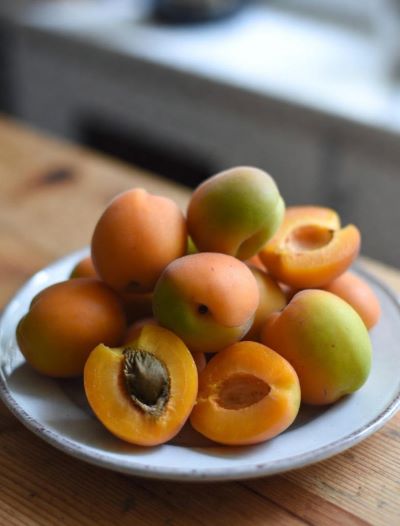 A white ceramic plate piled with fresh early-season apricots, some whole and some halved to reveal their bright orange flesh and pits. The apricots range from golden-yellow to soft orange with a delicate blush, resting on a rustic wooden table bathed in gentle natural light.