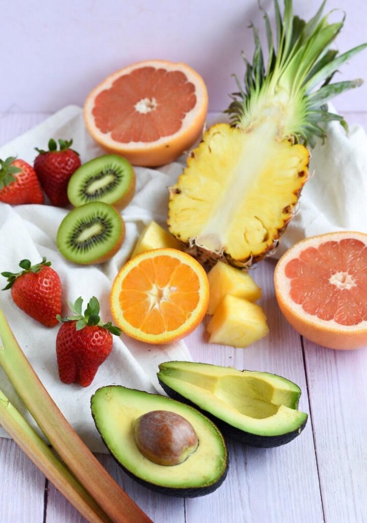 A colorful flat-lay arrangement of fresh early spring fruits in season, on a white cloth and light pink surface. The spread features a halved pineapple with leafy top, pink grapefruit halves, sliced green kiwi, an orange half, whole red strawberries, long rhubarb stalks, and a halved avocado with the pit visible.