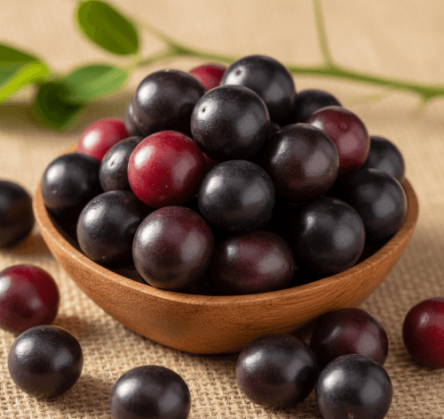 A close-up, studio photograph of a small wooden bowl filled with round, ripe berries, likely Karonda (Carissa carandas). The berries are a mix of deep reddish-purple and near-black colors. The bowl is sitting on a coarse, light-brown burlap surface. Several berries have spilled out and are scattered around the bowl. In the blurred background, a small branch with green leaves is visible. The lighting is focused, highlighting the glossy surface and rich color of the berries.